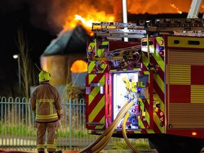 Obrazek przedstawia strażaka w pracy, w tle palący się budynek