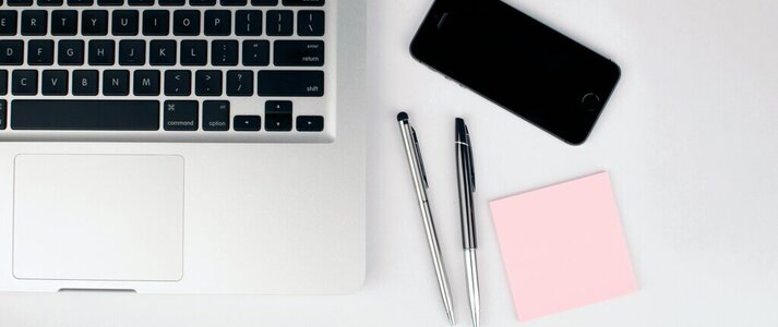 Obrazek przedstawia laptopa, telefon, dwa długopisy i karteczki różowe.